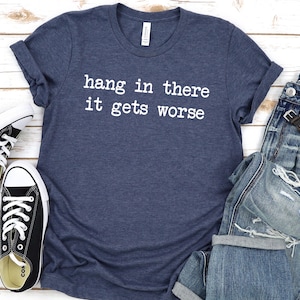 May include: A navy blue t-shirt with the text "hang in there it gets worse" in white, paired with distressed blue jeans, a wristwatch, and black and white sneakers. The items are arranged on a light wood-grain surface.