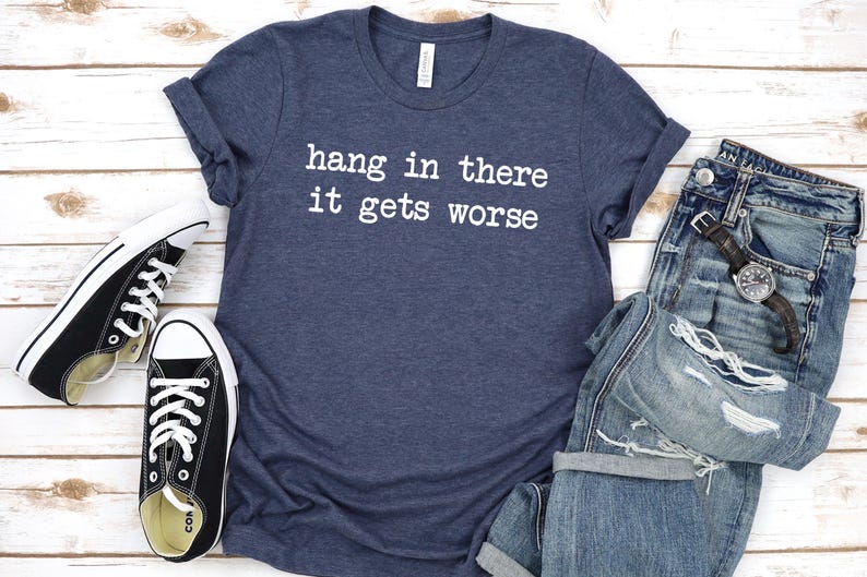 May include: A navy blue t-shirt with the text "hang in there it gets worse" in white, paired with distressed blue jeans, a wristwatch, and black and white sneakers. The items are arranged on a light wood-grain surface.