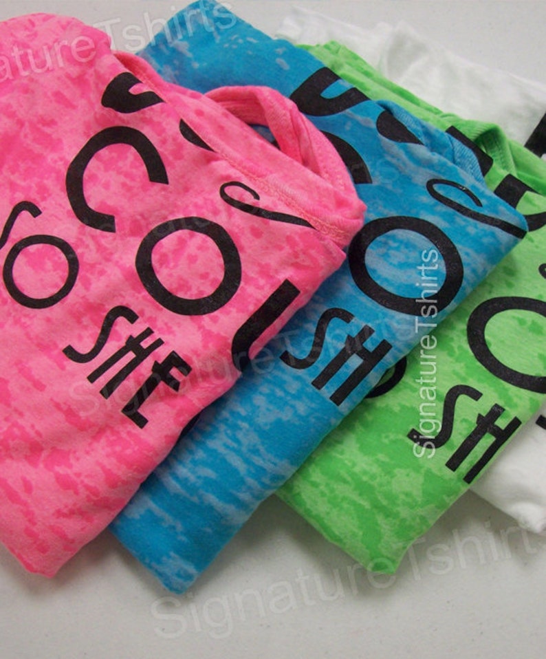 May include: Four t-shirts in different colors with the text "She's So Cool" printed on them. The shirts are stacked on top of each other, with the pink shirt on top and the white shirt on the bottom.