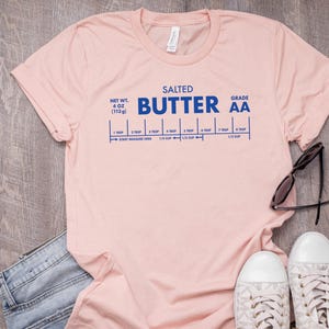 May include: A pink t-shirt with a blue graphic that reads "SALTED BUTTER AA" and includes a measurement guide for tablespoons and cups. The shirt is laid flat on a woodgrain surface.
