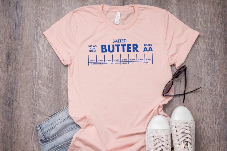 May include: A pink t-shirt with a blue graphic that reads "SALTED BUTTER AA" and includes a measurement guide for tablespoons and cups. The shirt is laid flat on a woodgrain surface.
