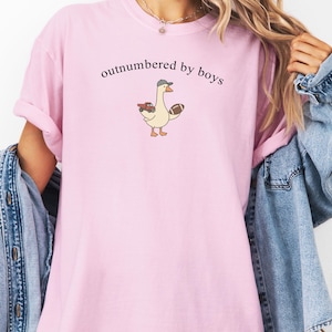 May include: A pale pink t-shirt with the words "outnumbered by boys" printed in black. The shirt features a cartoon goose wearing a hat and holding a football and a toy car. The sleeves are rolled up.