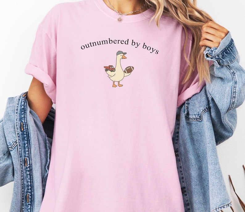 May include: A pale pink t-shirt with the words "outnumbered by boys" printed in black. The shirt features a cartoon goose wearing a hat and holding a football and a toy car. The sleeves are rolled up.