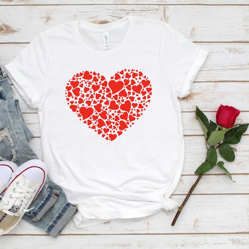 K&ouml;nnte beinhalten: Wei&szlig;es T-Shirt mit einem roten Herz aus kleineren roten Herzen. Das Shirt liegt flach auf einer wei&szlig;en Holzoberfl&auml;che mit einer blauen Jeans und einer roten Rose.