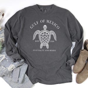 Op de afbeelding: Een grijs langarm T-shirt met een witte afbeelding van een zeeschildpad en de tekst "Gulf of Mexico Est. 1550 Stay Salty and Resist".