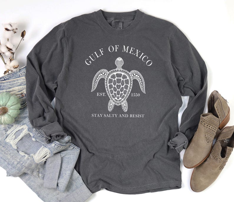 Op de afbeelding: Een grijs langarm T-shirt met een witte afbeelding van een zeeschildpad en de tekst "Gulf of Mexico Est. 1550 Stay Salty and Resist".