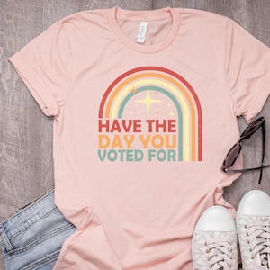 May include: A light pink t-shirt with a rainbow graphic and the text "Have the day you voted for." The shirt is paired with blue jeans, white sneakers, and sunglasses. The shirt is made of a soft material.