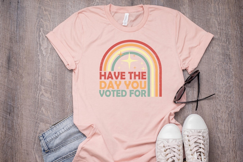 May include: A light pink t-shirt with a rainbow graphic and the text "Have the day you voted for." The shirt is paired with blue jeans, white sneakers, and sunglasses. The shirt is made of a soft material.