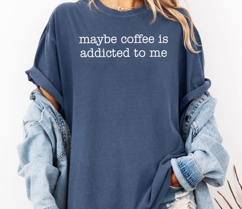 May include: A navy blue t-shirt with the white text "maybe coffee is addicted to me." The shirt is worn with a light blue denim jacket. The sleeves of the shirt are rolled up.
