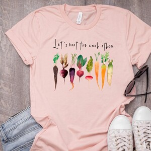 May include: Pink t-shirt with the text "Let's root for each other" and a watercolor illustration of various vegetables, including carrots, beets, and radishes.