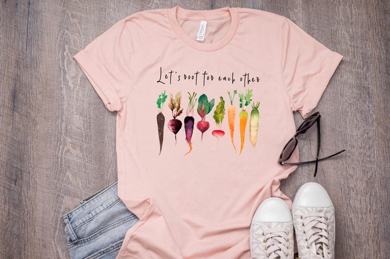 May include: Pink t-shirt with the text "Let's root for each other" and a watercolor illustration of various vegetables, including carrots, beets, and radishes.