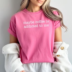 May include: A bright pink t-shirt with the phrase "maybe coffee is addicted to me" printed in white. The shirt has rolled-up sleeves and is paired with a white cardigan and gray sweatpants.