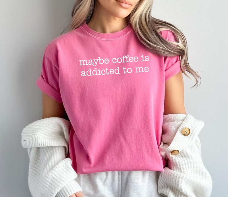 May include: A bright pink t-shirt with the phrase "maybe coffee is addicted to me" printed in white. The shirt has rolled-up sleeves and is paired with a white cardigan and gray sweatpants.