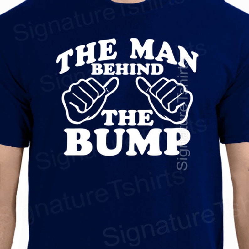 May include: A navy blue t-shirt with white text that reads "The Man Behind The Bump". The text is surrounded by two white thumbs up.