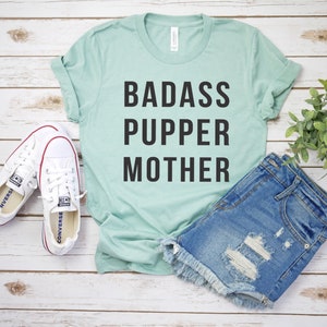 May include: A light blue t-shirt with the text "BADASS PUPPER MOTHER" in black lettering. The shirt is laid flat on a white wooden surface with a pair of white sneakers and a pair of denim shorts.