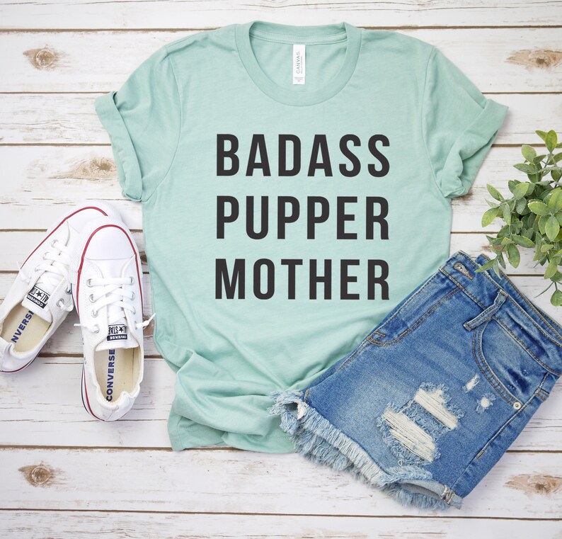 May include: A light blue t-shirt with the text "BADASS PUPPER MOTHER" in black lettering. The shirt is laid flat on a white wooden surface with a pair of white sneakers and a pair of denim shorts.