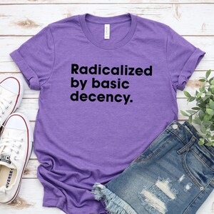 May include: A purple t-shirt with the text "Radicalized by basic decency" in black. The shirt is paired with denim shorts and white sneakers. The shirt is made of a soft material.
