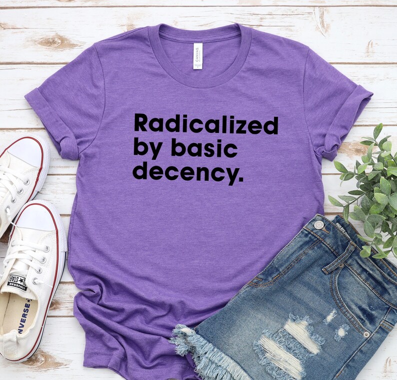 May include: A purple t-shirt with the text "Radicalized by basic decency" in black. The shirt is paired with denim shorts and white sneakers. The shirt is made of a soft material.