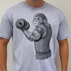 May include: A gray t-shirt with a black and white illustration of a muscular monkey lifting a dumbbell. The monkey is wearing a sleeveless shirt and has a banana in its hand. The text "Signature Tshirts" is printed on the shirt.