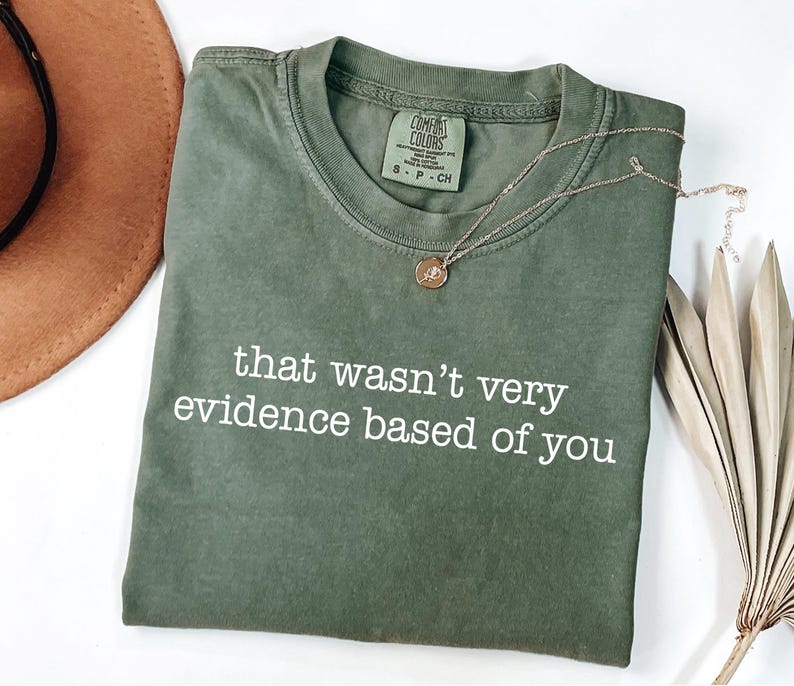 May include: An olive green Comfort Colors t-shirt with the text "that wasn't very evidence based of you" in white. A gold necklace with a pendant and a brown hat are also in the image.