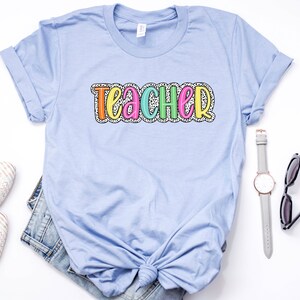 Puede incluir: Una camiseta azul claro con la palabra "teacher" escrita en letras de colores con un patr&oacute;n de lunares blancos.