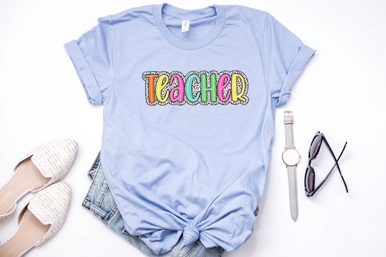 Puede incluir: Una camiseta azul claro con la palabra "teacher" escrita en letras de colores con un patr&oacute;n de lunares blancos.
