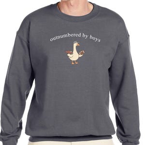 May include: Dark gray sweatshirt with the phrase "outnumbered by boys" above a cartoon goose wearing a baseball cap and holding a football and a toy truck. The sweatshirt is a crew neck style.