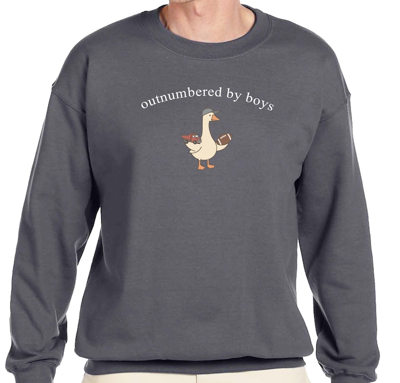 May include: Dark gray sweatshirt with the phrase "outnumbered by boys" above a cartoon goose wearing a baseball cap and holding a football and a toy truck. The sweatshirt is a crew neck style.