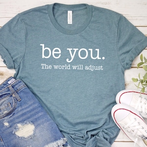 Puede incluir: Una camiseta de color verde azulado con las palabras "be you. The world will adjust" en letras blancas. La camiseta se combina con pantalones cortos de mezclilla y zapatillas blancas con detalles en rojo, todo sobre una superficie de madera clara.