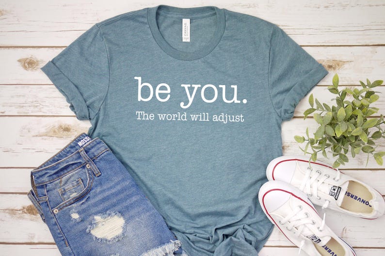 Puede incluir: Una camiseta de color verde azulado con las palabras "be you. The world will adjust" en letras blancas. La camiseta se combina con pantalones cortos de mezclilla y zapatillas blancas con detalles en rojo, todo sobre una superficie de madera clara.