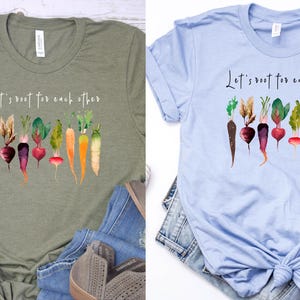 May include: Two t-shirts with a green and a light blue background. Both shirts feature a watercolor illustration of vegetables with the text "Let's root for each other".