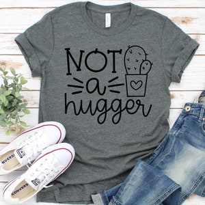 May include: A heather gray t-shirt with the text "NOT a hugger" and a cactus graphic. The shirt is laid flat on a white wood background with a pair of white sneakers and blue jeans.