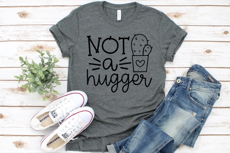 May include: A heather gray t-shirt with the text "NOT a hugger" and a cactus graphic. The shirt is laid flat on a white wood background with a pair of white sneakers and blue jeans.