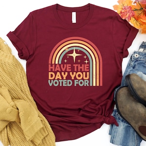 May include: A maroon t-shirt with a retro rainbow design and the text "HAVE THE DAY YOU VOTED FOR". The graphic includes a starburst and star accents. The shirt is styled with jeans and boots.