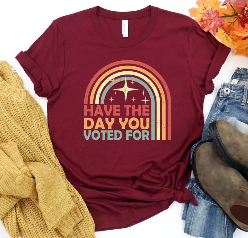 May include: A maroon t-shirt with a retro rainbow design and the text "HAVE THE DAY YOU VOTED FOR". The graphic includes a starburst and star accents. The shirt is styled with jeans and boots.