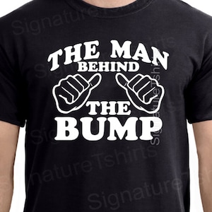 May include: Black t-shirt with white text that reads "The Man Behind The Bump". The text is surrounded by two white thumbs up.
