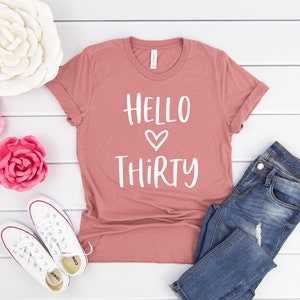 May include: A dusty rose colored t-shirt with the text "HELLO THIRTY" in white with a heart symbol between the words.