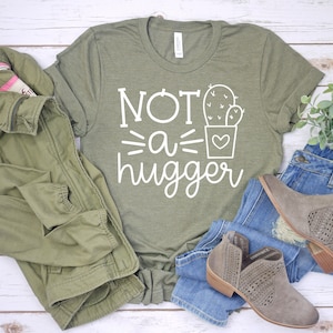 May include: A green t-shirt with white text that reads "Not a hugger" and a cactus graphic. The shirt is laid out on a white wooden surface with a green jacket, blue jeans, and brown boots.
