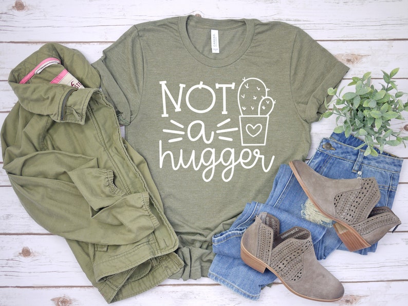 May include: A green t-shirt with white text that reads "Not a hugger" and a cactus graphic. The shirt is laid out on a white wooden surface with a green jacket, blue jeans, and brown boots.