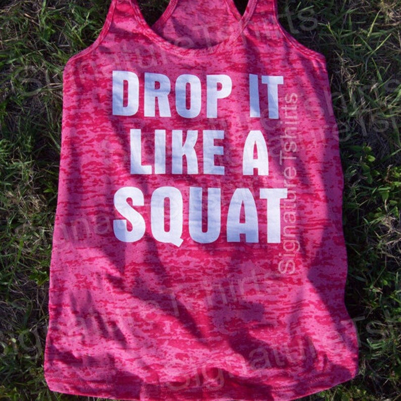 Może przedstawiać: R&oacute;żowa koszulka na ramiączkach z białym, przetartym nadrukiem, na kt&oacute;rym widnieje napis "Drop it like a squat".