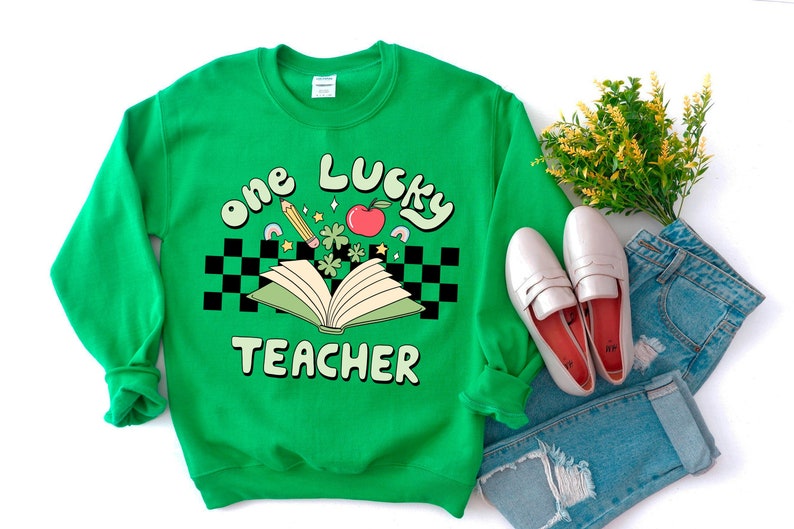 May include: Green sweatshirt with the text "one lucky teacher" in a retro font. The design includes a book, a pencil, a red apple, and four-leaf clovers.