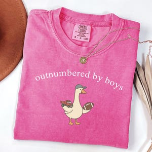 May include: A vibrant pink t-shirt with the words "outnumbered by boys" in white. The shirt features a cartoon goose wearing a baseball cap and holding a football and a toy truck. A gold necklace is draped across the shirt.