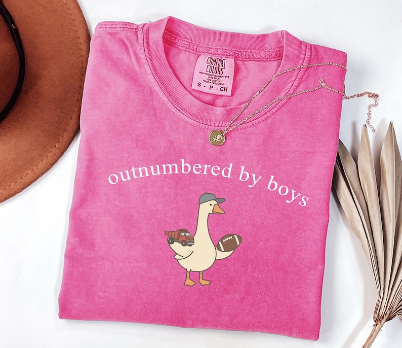 May include: A vibrant pink t-shirt with the words "outnumbered by boys" in white. The shirt features a cartoon goose wearing a baseball cap and holding a football and a toy truck. A gold necklace is draped across the shirt.