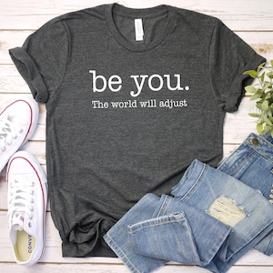 Puede incluir: Una camiseta gris oscuro con la frase blanca "be you. The world will adjust". Se combina con vaqueros de mezclilla azul claro y zapatillas de lona blancas con detalles rojos.