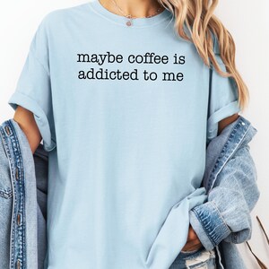 May include: Light blue t-shirt with the text "maybe coffee is addicted to me" in black. The shirt has short sleeves and a relaxed fit. A denim jacket is draped over the shoulders.