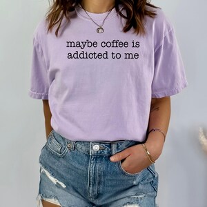 May include: A light purple t-shirt with the phrase "maybe coffee is addicted to me" printed in black. The shirt is a casual style, with short sleeves and a relaxed fit. The model is wearing denim shorts.