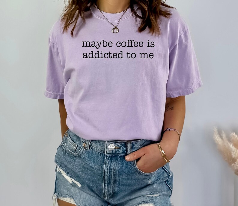 May include: A light purple t-shirt with the phrase "maybe coffee is addicted to me" printed in black. The shirt is a casual style, with short sleeves and a relaxed fit. The model is wearing denim shorts.