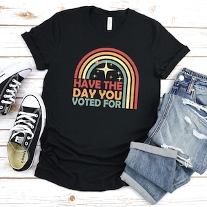 May include: Black t-shirt with a rainbow graphic and the text "HAVE THE DAY YOU VOTED FOR". The design features a retro color palette. A pair of black and white sneakers and denim shorts are next to the shirt.