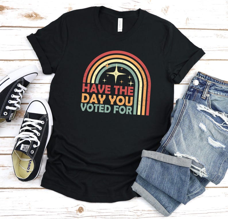 May include: Black t-shirt with a rainbow graphic and the text "HAVE THE DAY YOU VOTED FOR". The design features a retro color palette. A pair of black and white sneakers and denim shorts are next to the shirt.