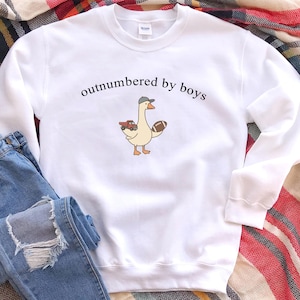 May include: White sweatshirt with the text "outnumbered by boys" above a cartoon goose wearing a hat and holding a football and a toy car. The sweatshirt is paired with blue jeans on a plaid blanket.